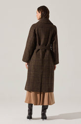A woman is wearing a long, dark brown plaid coat with a belt at the waist and a wide collar. The coat has a classic checkered pattern and falls below the knees. Underneath, she is wearing a beige or tan pleated skirt that reaches mid-calf. She is also wearing tall black heeled boots. Her hair is dark brown, styled straight, and she is wearing gold earrings.